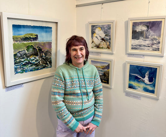 A photograph of Orkney artist Jane Glue standing in front of a few of her paintings from her exhibition called 'It's all about the light' at The Loft gallery in St Margarets Hope in Orkney.