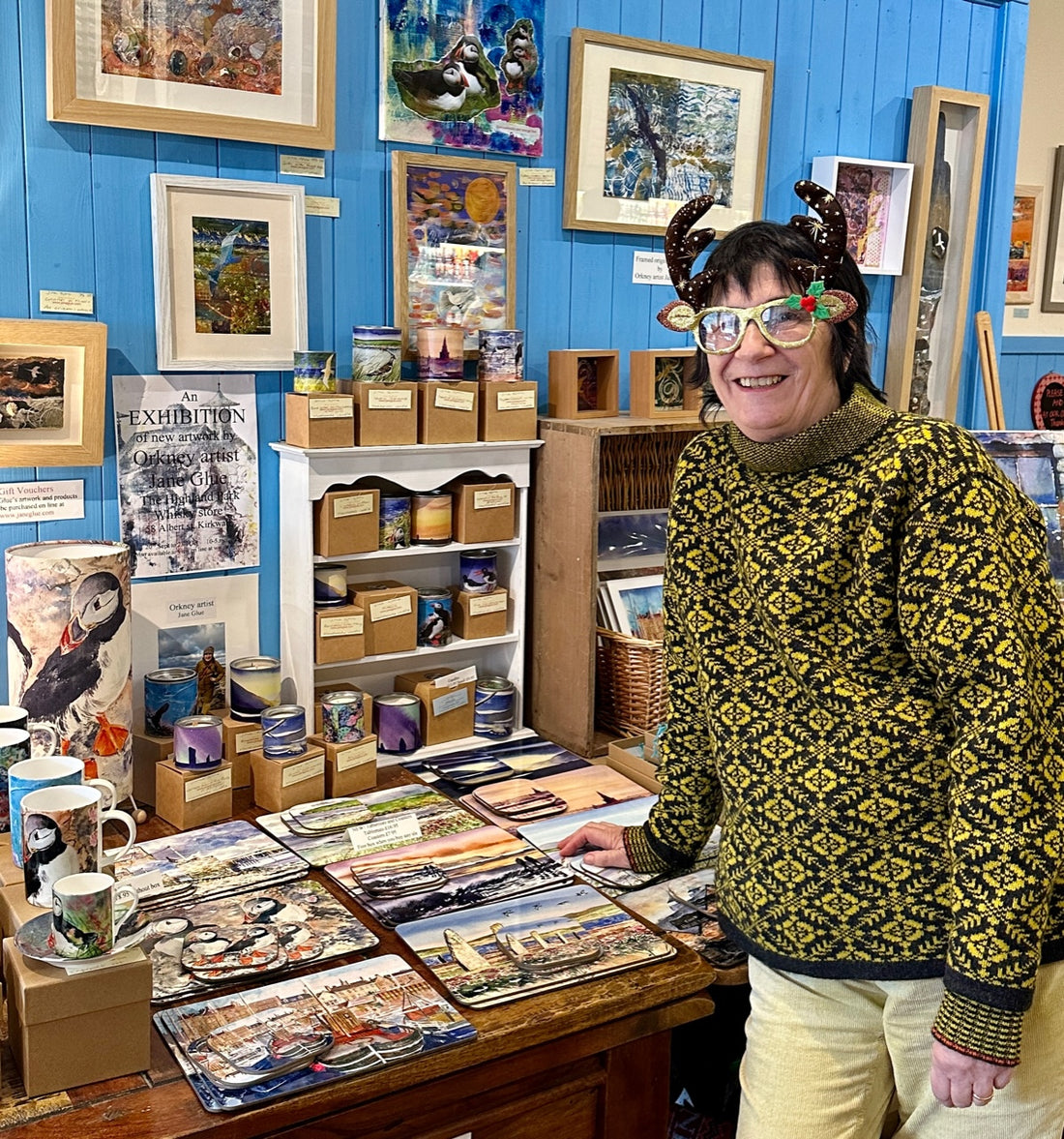 Orkney artist Jane Glue wearing Christmas glasses standing in front of her gifts table in her sister Judith Glue's shop in Kirkwall