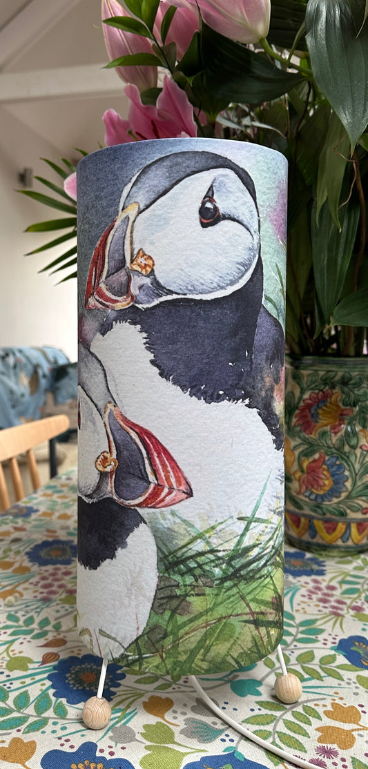 a tall table lamp with three wooden balls for feet printed with a watercolour image of two puffins sitting amongst green grass