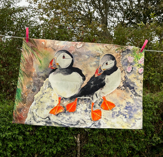 a linen t towel printed with a watercolour painting of two puffins with bright orange feet standing on a cliff edge