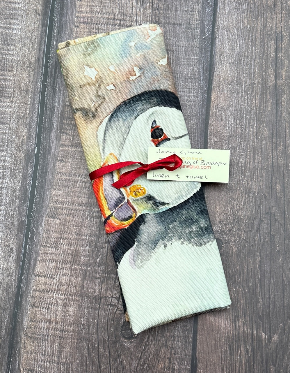 a linen t towel printed with a watercolour painting of two puffins with bright orange feet standing on a cliff edge