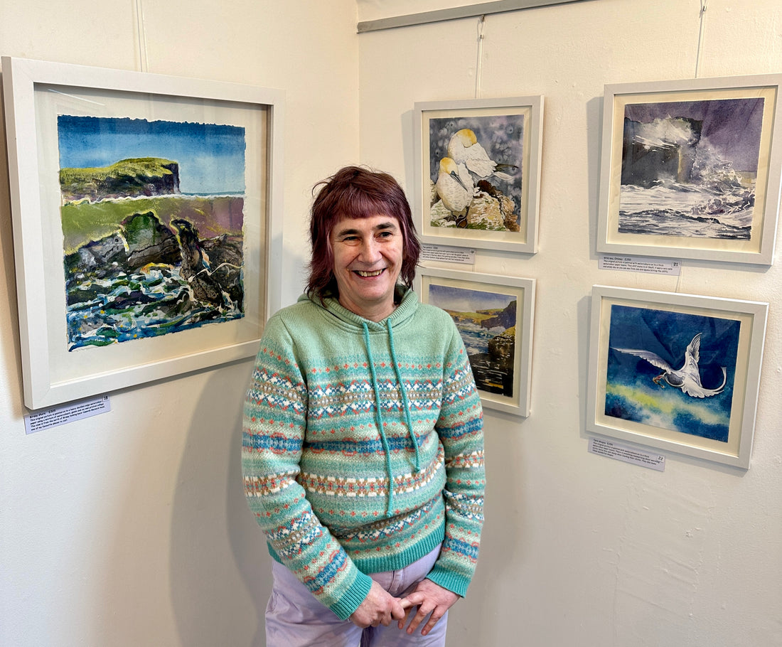 A photograph of Orkney artist Jane Glue standing in front of a few of her paintings from her exhibition called 'It's all about the light' at The Loft gallery in St Margarets Hope in Orkney.