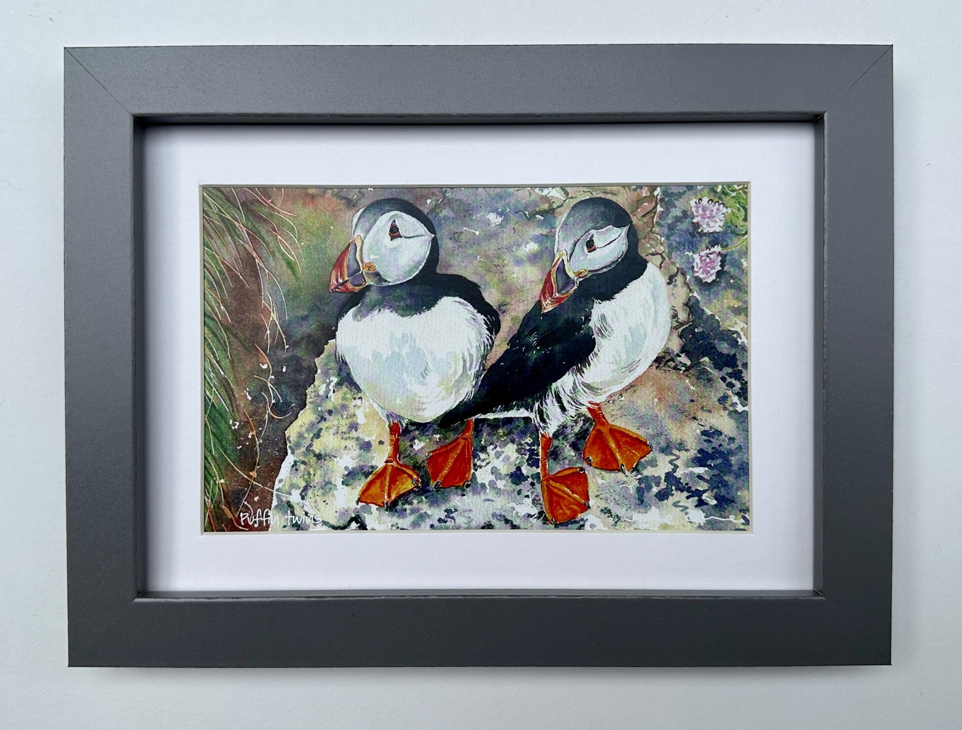 small framed print in a grey wood moulding and white mount of a watercolour painting  of two puffins standing on a cliff