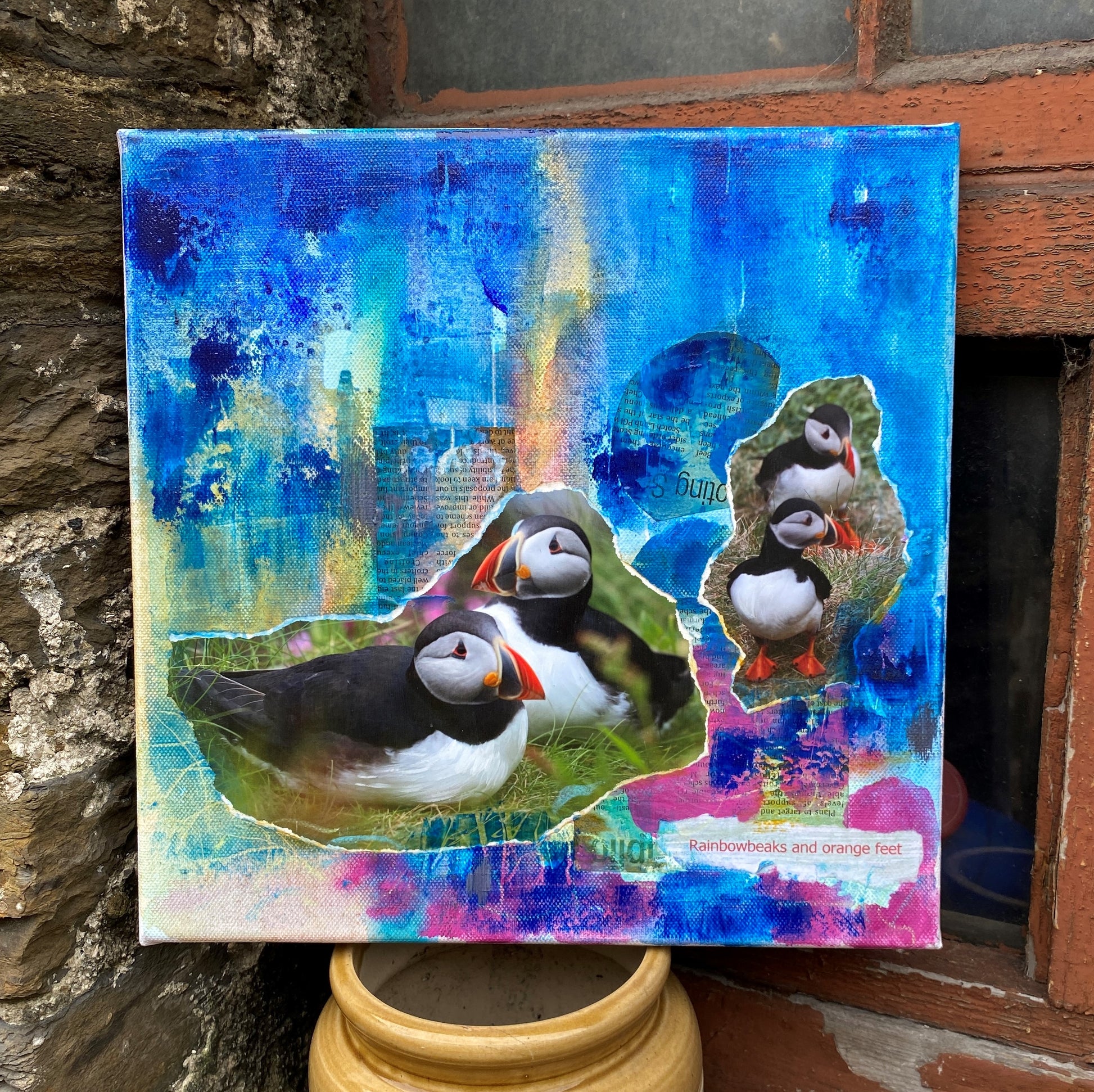 an original mixed media painting of four puffins standing and sitting on a grassy cliff with a blue and pink background