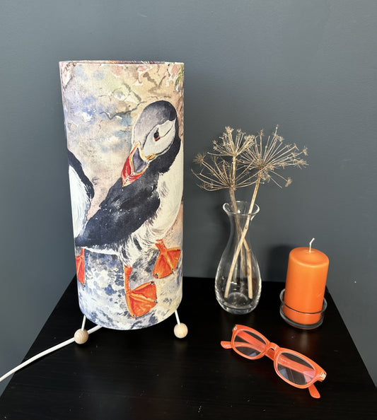 a tall table lamp with three wooden balls for feet printed with a watercolour image of two puffins standing on a cliff